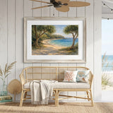 A framed coastal landscape painting hangs on a white wood-paneled wall above a woven rattan bench with pillows and a throw blanket. The scene includes a porch ceiling fan, string lights, a rug, and an ocean view visible through an open doorway.