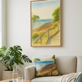 A framed coastal landscape painting hangs on a light wall above a beige sofa with a matching printed pillow, in a styled living room with a side table, plant, and blue chair.