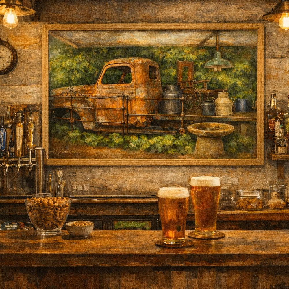 A framed art print depicting an old rusted pickup truck in a green field, displayed on a rustic bar wall above a wooden counter with two glasses of beer in a cozy, warmly lit pub setting.