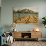 A framed landscape painting of a vineyard hangar and rows of grapevines, displayed on a sage green wall in a modern living room setting with a teal armchair, wooden console, and potted plant.