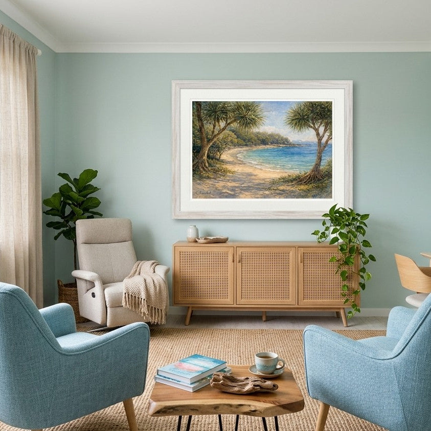 A framed landscape art print depicting a serene beach scene with trees and blue water, displayed on a light blue wall in a styled living room with two blue armchairs, a wooden cabinet, and a window view.