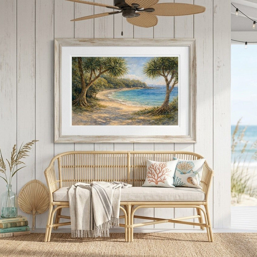 A framed coastal landscape painting hangs on a white wood-paneled wall above a woven rattan bench with pillows and a throw blanket. The scene includes a porch ceiling fan, string lights, a rug, and an ocean view visible through an open doorway.
