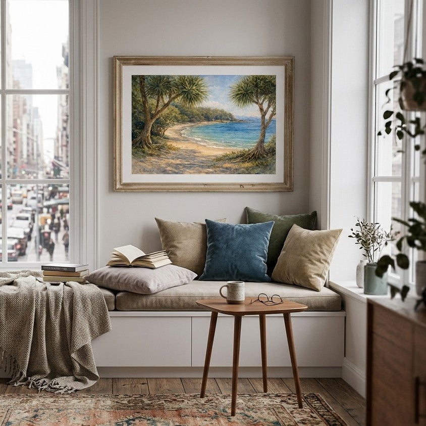 A framed landscape painting of a tree-lined beach hangs on a white wall above a cozy window seat with beige and blue pillows, a throw blanket, and a small wooden side table in a bright city apartment.
