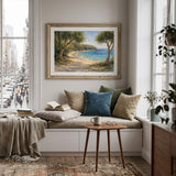 A framed landscape painting of a tree-lined beach hangs on a white wall above a cozy window seat with beige and blue pillows, a throw blanket, and a small wooden side table in a bright city apartment.