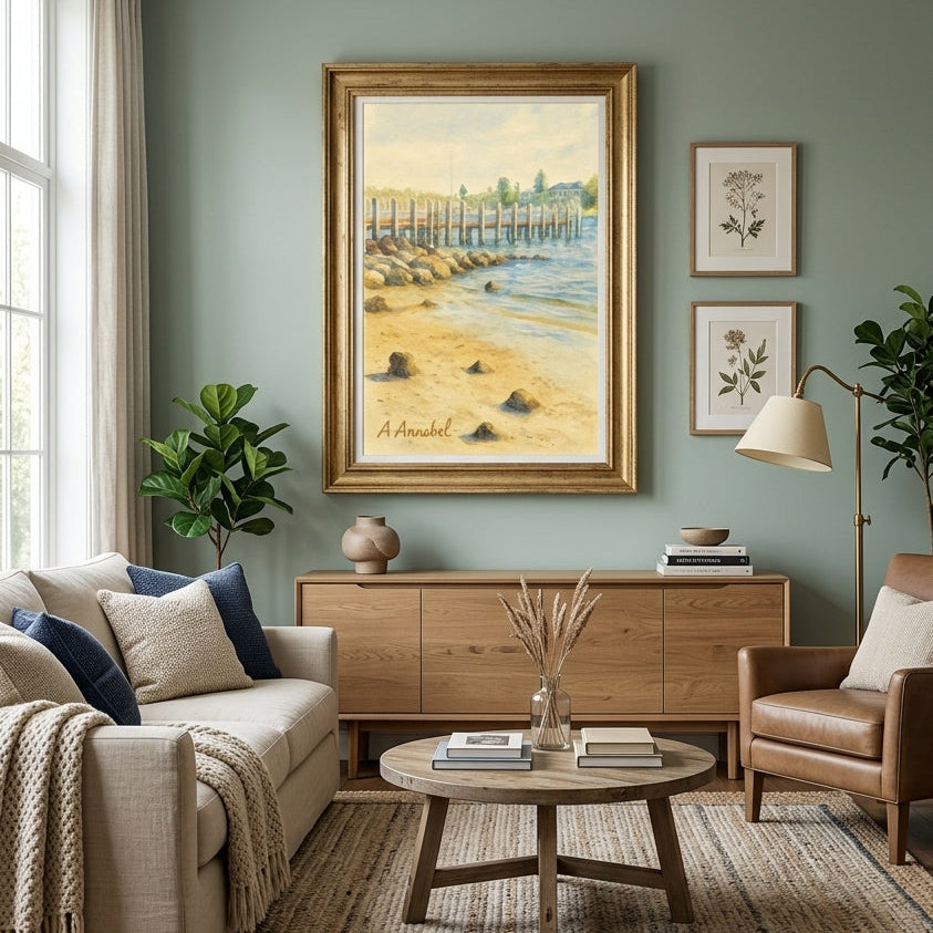 A framed art print depicting a sandy beach and wooden pier on the wall in a beautifully styled living room with a sage green wall, beige sofa, wooden coffee table, and brown leather armchair.