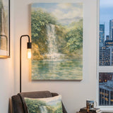 A vertical canvas painting of a scenic waterfall in a lush green forest, styled in a modern living room setting above a gray armchair with a matching pillow.