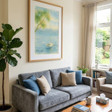 A framed landscape art print depicting a serene blue sea and green tree, hung on a cream wall above a gray fabric sofa in a styled living room.