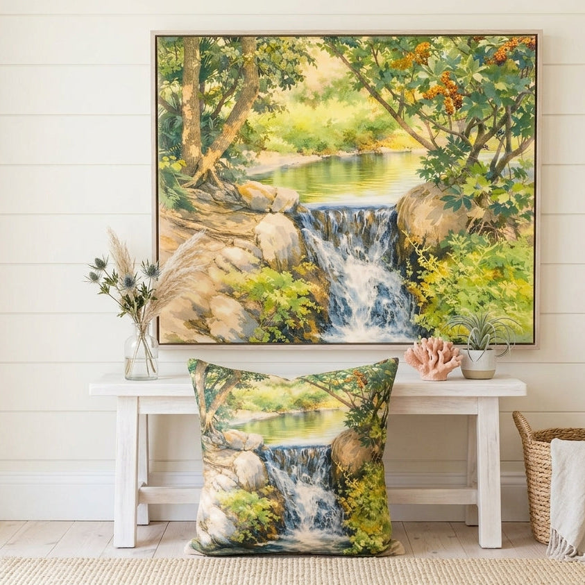 A framed landscape art print of a waterfall in a forest is displayed on a white wall above a white bench. A matching square throw pillow with the same print lies on the floor in front of the bench.