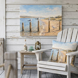 A coastal beach scene canvas print hangs on a white wood porch wall above a white Adirondack chair with a matching pillow, alongside a small wooden side table with decorative items.