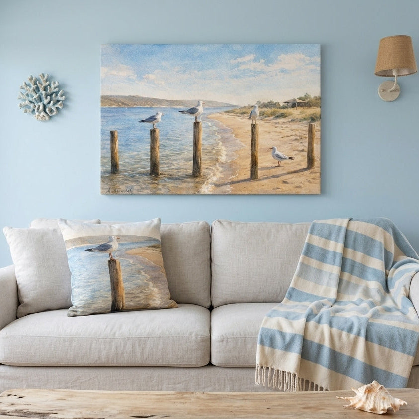 A coastal beach scene canvas print hanging on a light blue wall above a beige sofa with a blue and white striped throw blanket.