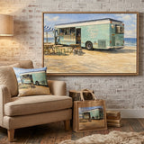 A framed art print depicting a vintage light blue beach food truck on a sandy shore, displayed on a white brick wall in a cozy living room setting with a beige armchair, lamp, and sofa.