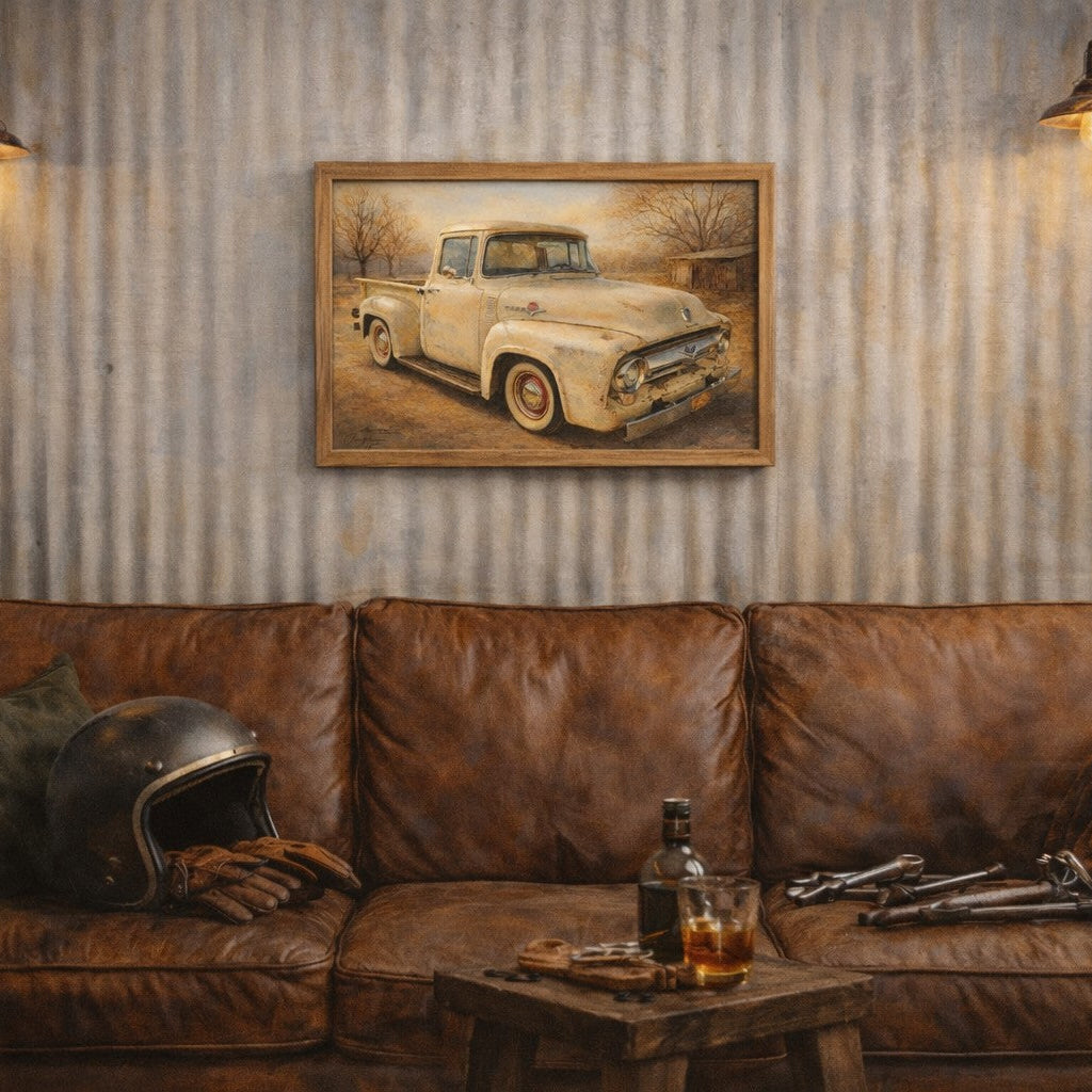 A framed vintage pickup truck painting hangs on a corrugated metal wall above a brown leather couch in a rustic room setting with two hanging lamps, a helmet, and a small table with a bottle and glass.