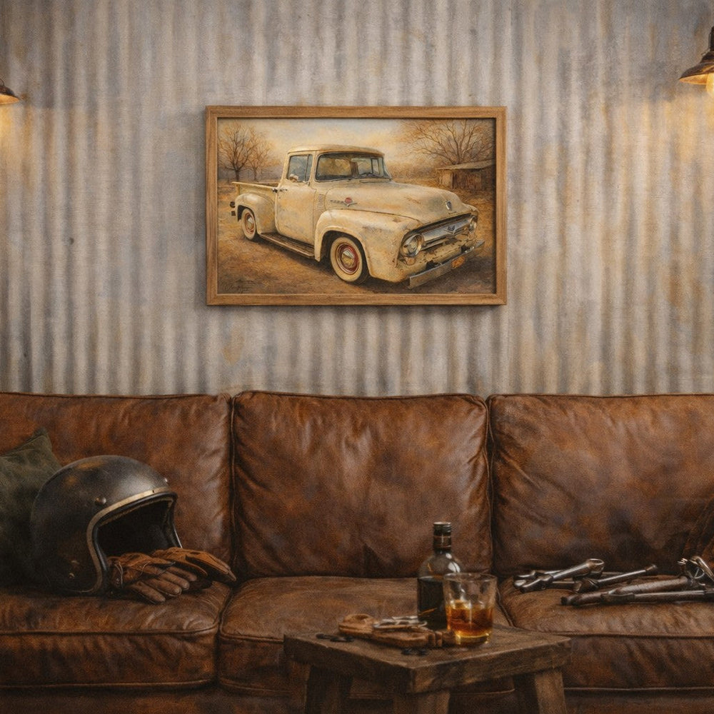 A framed vintage pickup truck painting hangs on a corrugated metal wall above a brown leather couch in a rustic room setting with two hanging lamps, a helmet, and a small table with a bottle and glass.